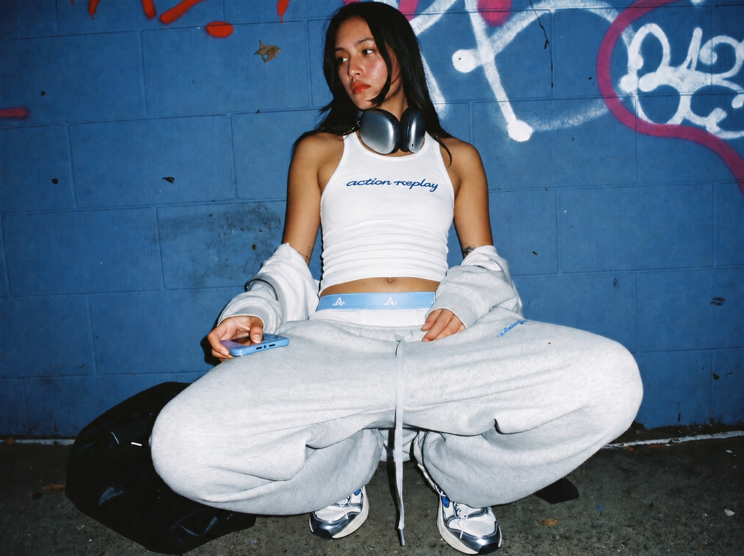 Model crouched against a blue wall wearing white Action Replay boxers layered under grey sweats.